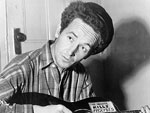 Photo, Woody Guthrie, half-length portrait, seated, facing front, playing a guitar that has a sticker attached reading: This Machine Kills Fascists, Mar. 8, 1943, Al Aumuller, Library of Congress