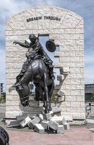 Statue of a person riding a horse through a white brick wall with the words “Breakin Through” on the top most part of the wall that is still intact.
