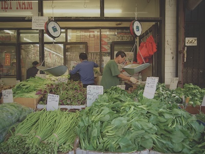 Crates of spinach, radishes, scallions, and other green vegetables sit in the foreground with prices. A few men stand behind the produce wearing gloves and interacting with the produce.