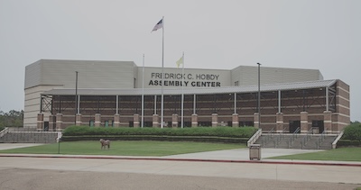 Outside of a large building that reads “Frederic C. Hobdy Assembly Center.” The building contains two flags, the US flag and a smaller yellow flag.