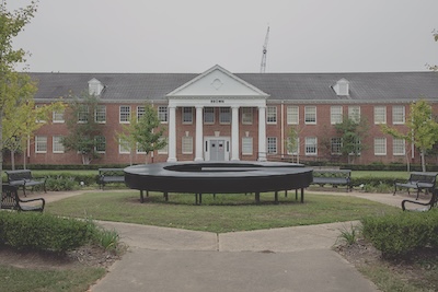 Outdoor setting with a brick building lining the back of the photograph. In the foreground, a large, horizontal sculpture of the letter G sits on a small patch of grass. The patch of grass is surrounded by a concrete path and a few benches.