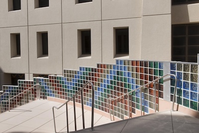 Outside photograph of a multi-colored glass wall descending alongside a cement stairwell. The glass wall takes up the bottom half of a building wall. The rest of the building’s wall is a light tan color.