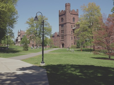 Outside in Mount Holyoke. A street light and paved path are in the front of the photograph. Next to those is a grassy field lined with trees, and a brick tower toward the back of the frame. In the distance, a few people are walking in the group