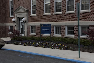 Brick building with rows of windows with a sign in front that reads “Potomac State College West Virginia University Science Hall”. The doorway is framed by a decorative cement design and two large white ball sconces. Between the building and the sidewalk there are shrugs and a flower bed.
