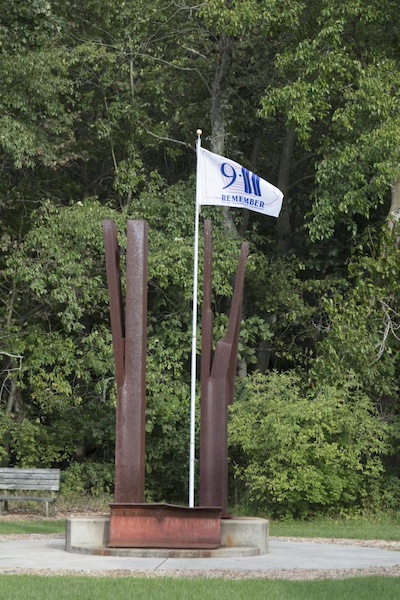 Two rusted steel beams like those from the Twin Tower rubble rise and each branch off to create four total branches. In the center, a white flag flies that reads &ldquo;9/11 Remember New York City Washington DC Pennsylvania&rdquo; with the outline of the Pentagon in the background