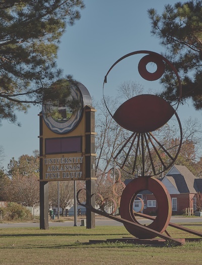 Outside image of a sign and a sculpture. The sign has two posts and a rounded top and reads University of Arkansas Pine Bluff. The sculpture is a bronze color with three abstract shapes stacked on top of one another.