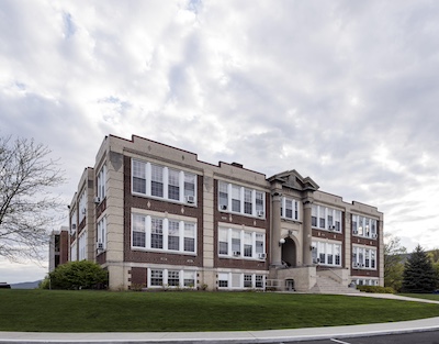 Large three story rectangular brick building with tall windows, stairs leading to the entrance, and two large columns by the door.