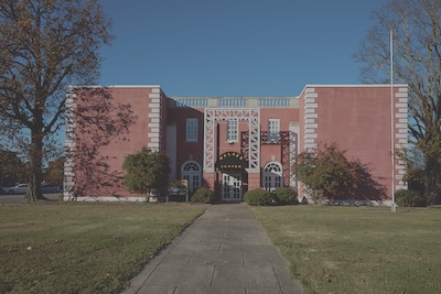 Outside of a large red brick building, which sits on a field of grass and a paved sidewalk leads up to the front door. Trees border the building.