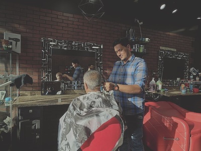 A barber cuts a gray-haired man’s hair in front of a mirror with an ornate border.