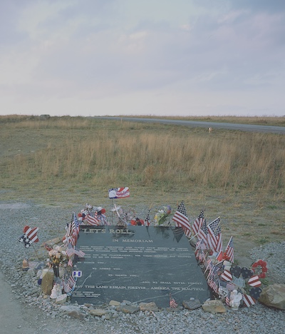 Memorial resembling a gravestone to honor the passengers on Flight 93. It is surrounded with flowers, small American flags and other items. The memorial appears to be near a wide open field and accompanies a larger museum.